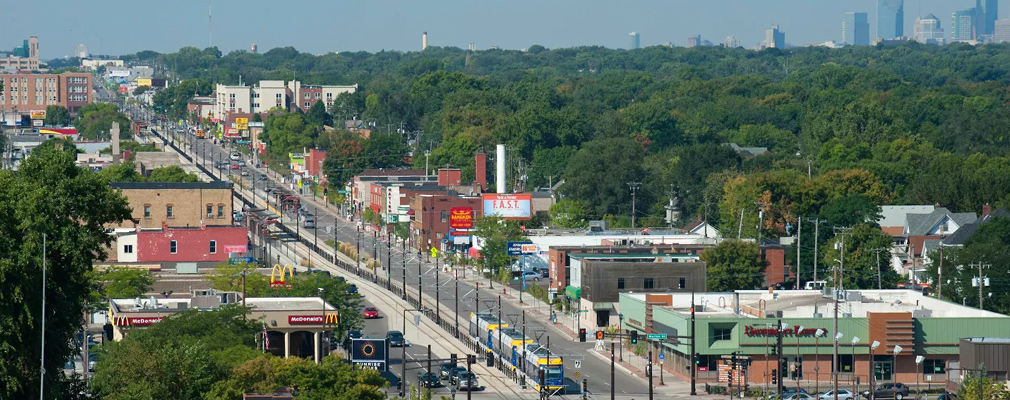 Aerial view of buildings, a main thoroughfare, and a light rail line and train running along the middle of the street.