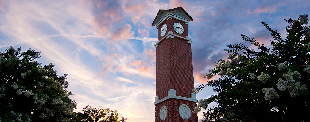 Photograph taken from ground level looking up at the Winston-Salem State University clock tower at dusk. The tower is made of brick highlighted with light-colored stone, and two of its clock faces are shown. Trees are planted around the tower.