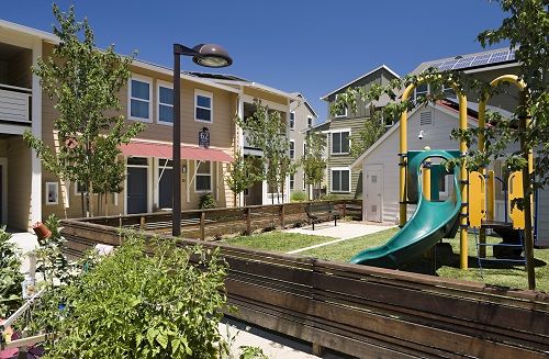 Photograph of a playground surrounded by a fence, small trees and shrubs, and low-rise apartments buildings.