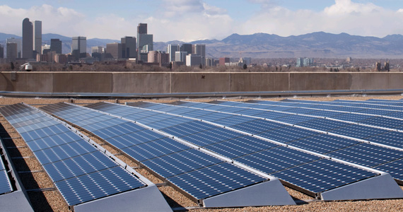 Image featuring several rows of solar panels on the rooftop of a building in Denver, Colorado. The Denver skyline, including skyscrapers and mountains, is visible in the background.