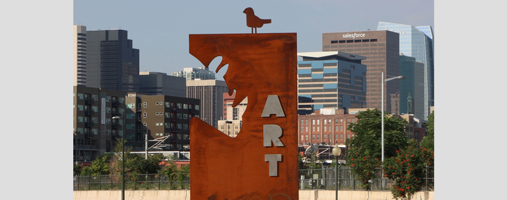 Photograph of an upright iron plate with carvings of a rhinoceros head and a bird and the word “ART” and with the skyline of downtown Denver in the background. 
