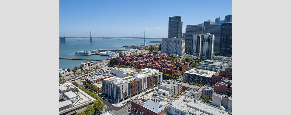 An aerial photograph of an urban development with low-rise buildings in the foreground and skyscrapers, San Francisco Bay, and a bridge in the background. 
