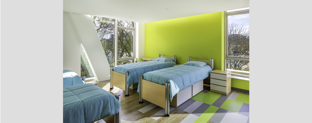 Four single beds in a brightly colored bedroom with windows providing natural light.