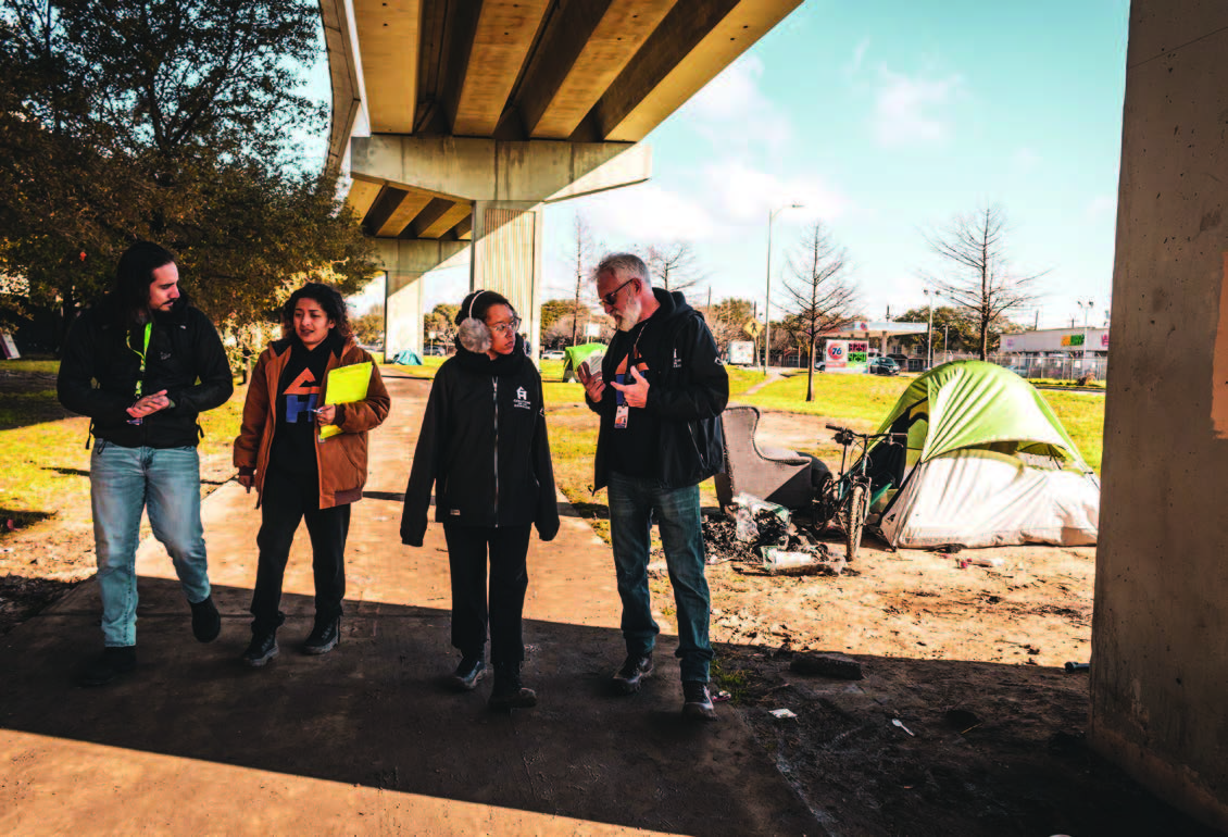 Four people walking under a concrete overpass with a tent on one side of the pathway.