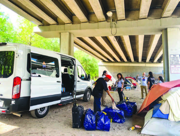 A van parked under a concrete overpass next to people and large blue plastic bags on the ground.