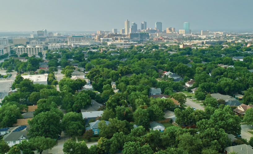 Aerial view of Dallas-Fort Worth area with residential areas with green trees in the foreground and multistory buildings in the background.