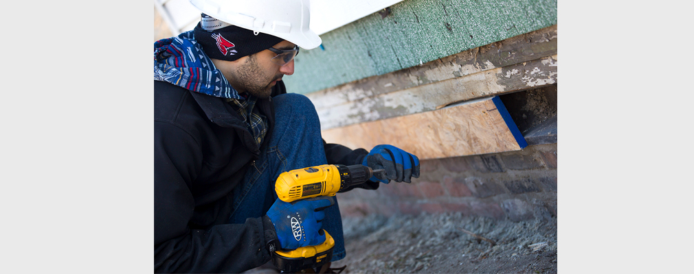 Photograph of a young man using an electric drill on the wood sheathing of a building.
