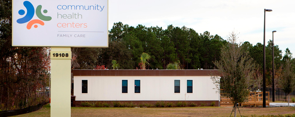 Photograph of a side façade of a modular building with a wooden accessibility ramp behind a sign which reads “Community Health Centers Family Care.” 