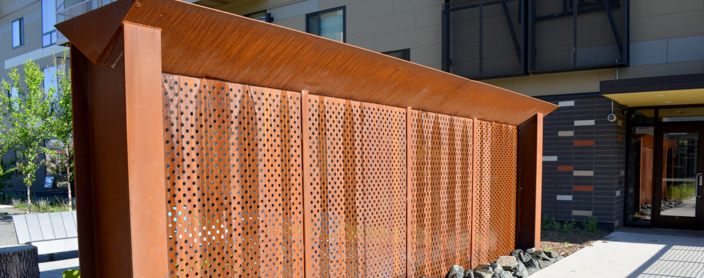 Photograph of a wall made of a perforated metal sheet, marked with vertical bands of condensed water, standing in a courtyard with a multistory residential building in the background.