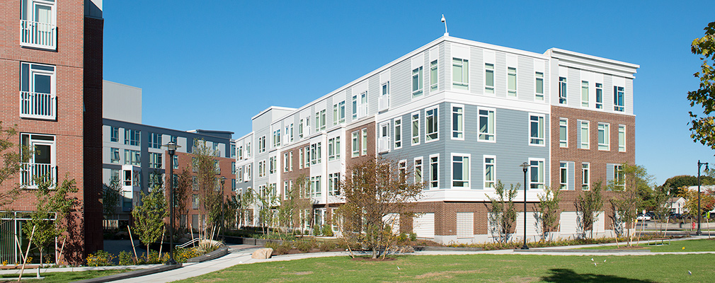 Photograph of two four-story apartment buildings on two sides of a landscaped courtyard and a 5-story mixed-use building at the rear of the courtyard.