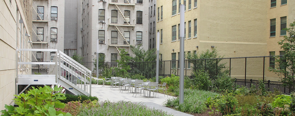 Photograph of a landscaped outdoor space beside the East Metro 99th Street building with a paved area accommodating several sets of tables and chairs.