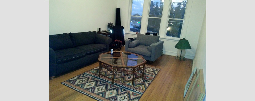 Photograph of a living room, with a table and an upholstered sofa and chair and with large windows on the far wall.