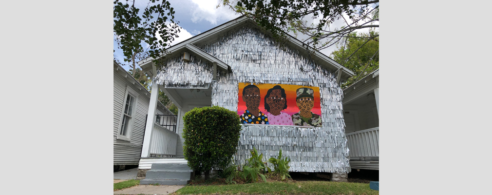 Photograph of the front façade of a shotgun house covered in metallic fringe and featuring a mural.
