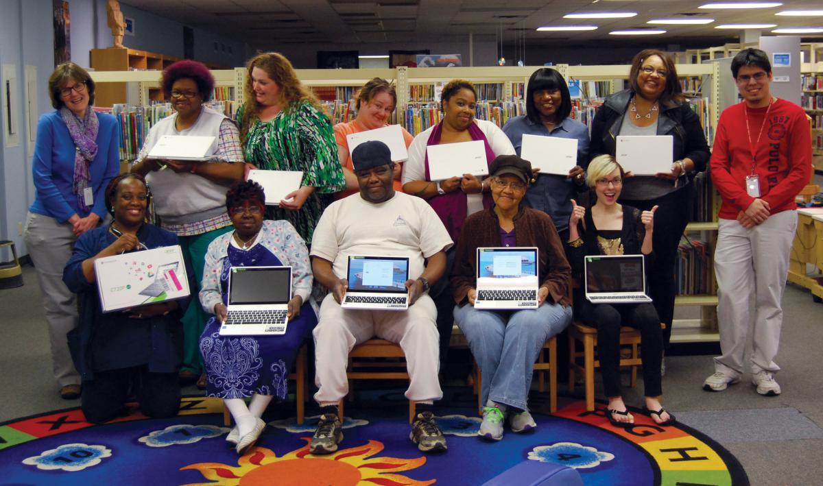 A group of men and women pose for the camera holding Chromebooks inside a library.