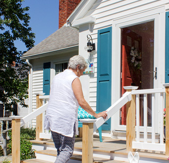 Photo shows a senior woman holding on to a railing and walking up stairs to the front stoop of a single-family home.