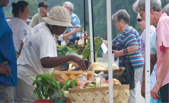Photo shows produce in baskets with buyers and sellers at a farmers market.