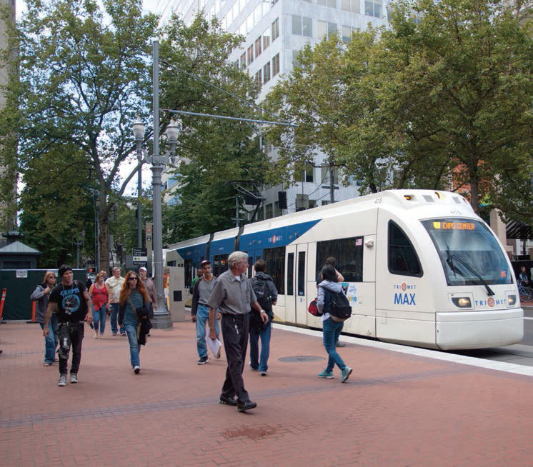 Photo shows people walking on a sidewalk with a light rail train stopped next to them.