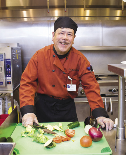 Photo shows a man holding a knife to cut vegetables on a cutting board in a hospital kitchen.