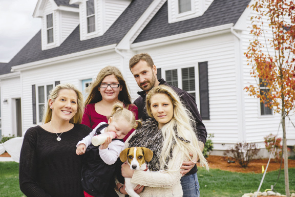 Photo shows a family of five and their dog posing in front of their home.