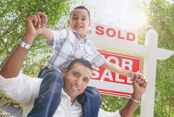 A Hispanic father carries his son on his shoulders while standing in front of a For Sale sign.
