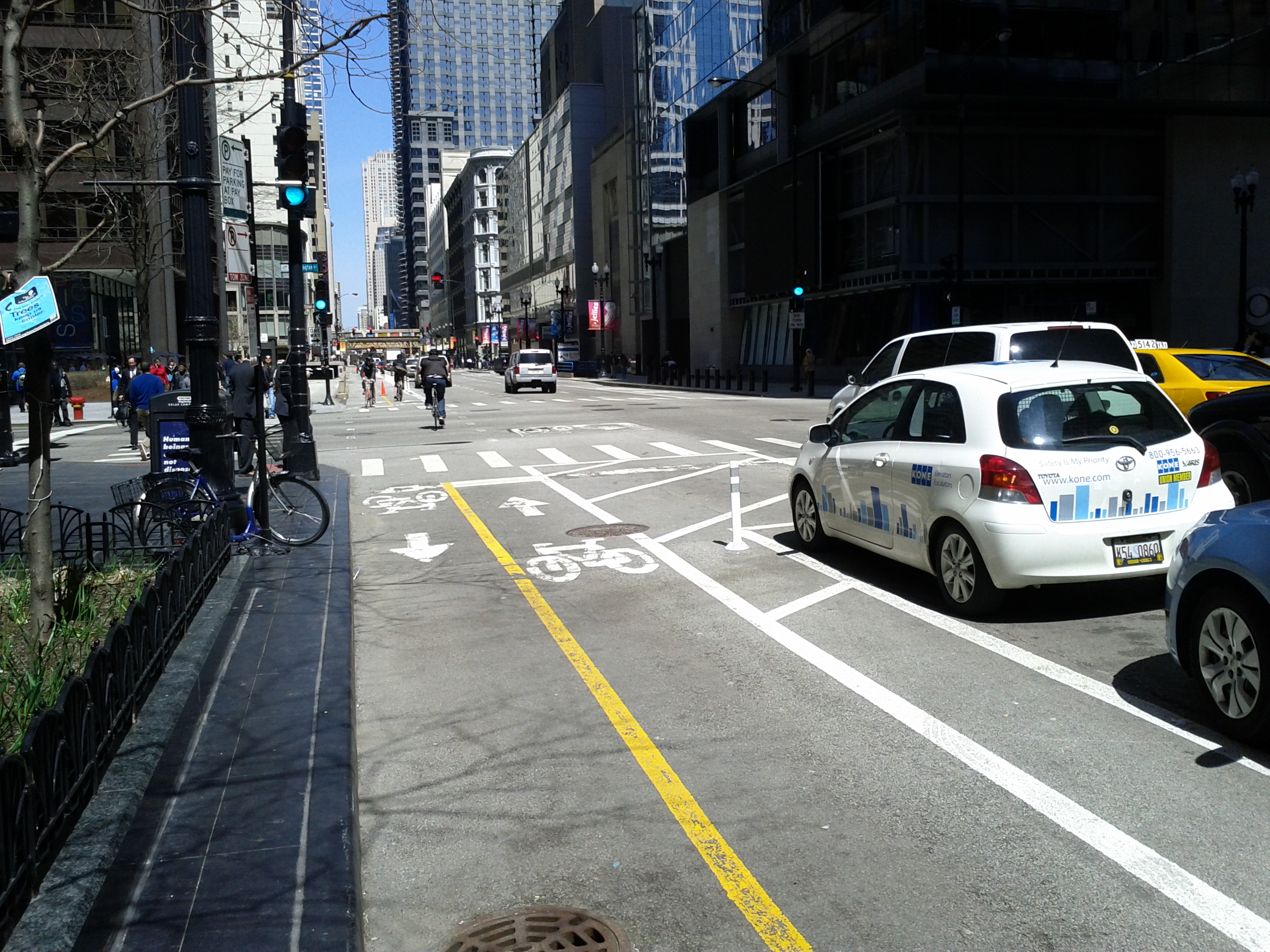 A photograph taken at street level in the two-way protected bike lane on Dearborn Street near its intersection with Washington Street) in downtown Chicago. Bicyclists are using the bike lane in the distance. Parked vehicles and a striped area separate the lane from moving traffic on the right of the photograph. On the left, the street curb and a planted area separate the bike lane from the sidewalk. 